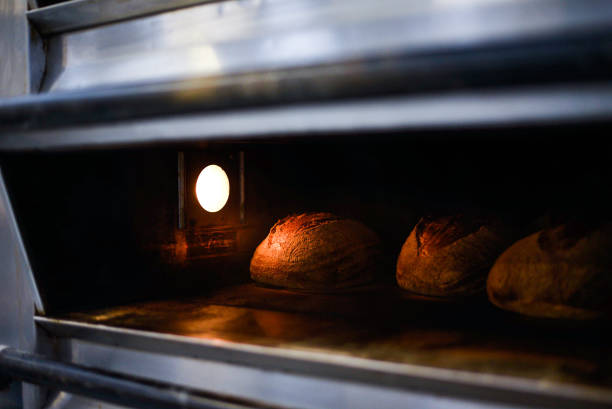 3 artisan sourdough loaves bake in a large commercial bakery oven, lit by the interior oven bulb. The loaves have completed the mallard process and will be removed from the oven within seconds. Real fresh bread.