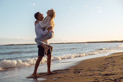 Full length photo of man holding laughing woman in his arms and looking at her while standing on sandy beach by the ocean