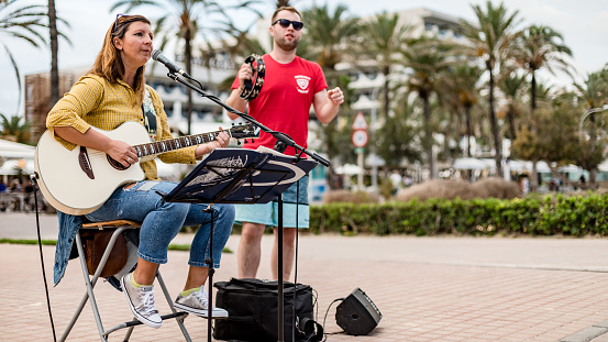 Palma de Majorca, Spain, Balearic Islands - October 14, 2018: People are street musicians on the beach. Man and woman playing guitar and singing microphone. Palms, Mediterranean sea around.