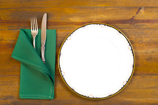 Place setting on a rustic wooden tabletop.  White china plate with dainty trim around edges. Green napkin with silver fork and knife.