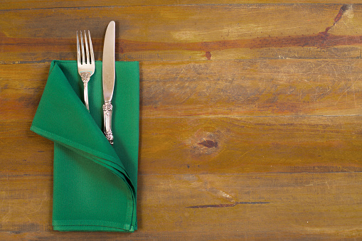 Place setting on a rustic wooden tabletop.   Green napkin with silver fork and knife.