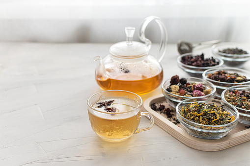A glass cup with hot tea and a kettle on a bright table by the window. Tray with different varieties of herbal tea on a white background. A delicious and healthy drink to strengthen immunity.