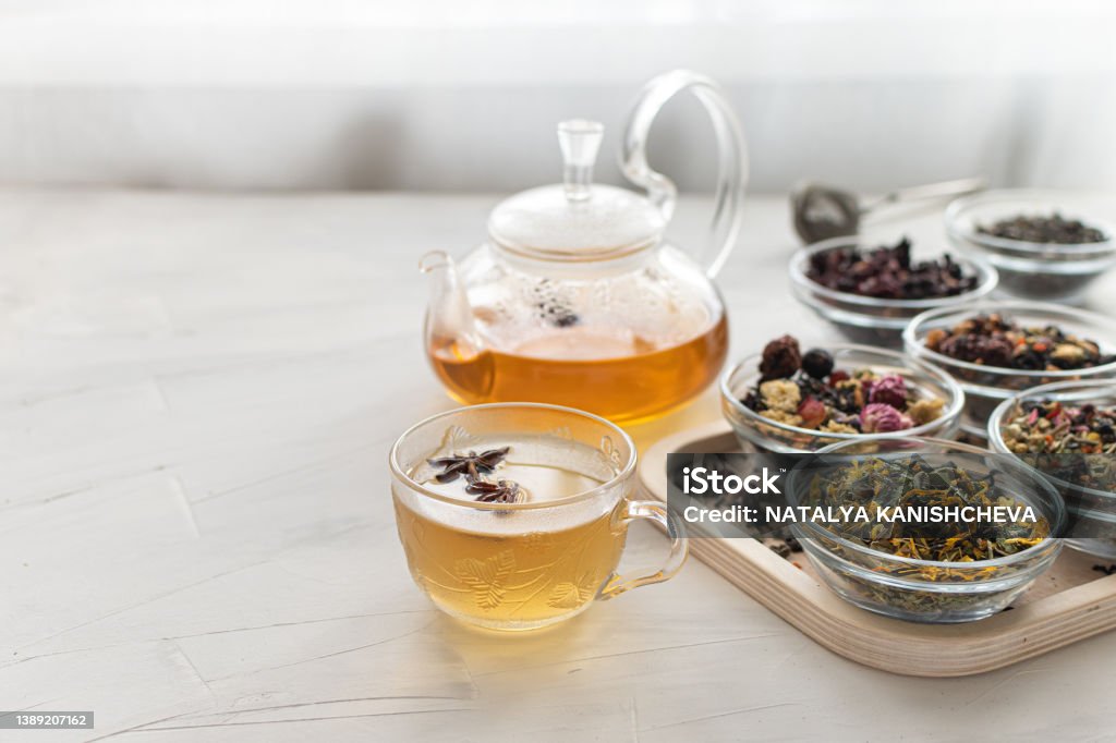 Glass cup with hot tea and a kettle on a bright table by the window Tray with different varieties of herbal tea on a white background Delicious and healthy drink to strengthen immunity Space for text A glass cup with hot tea and a kettle on a bright table by the window. Tray with different varieties of herbal tea on a white background. A delicious and healthy drink to strengthen immunity. Tea - Hot Drink Stock Photo