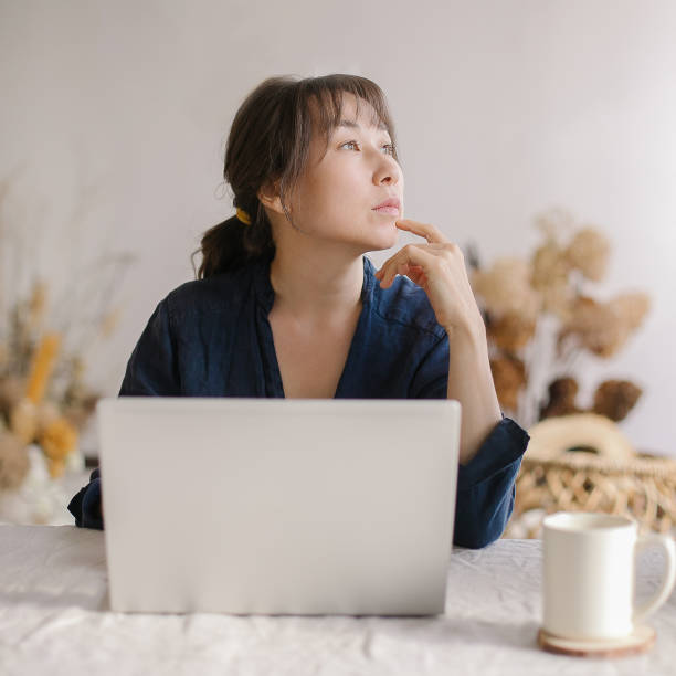 istock Thoughtful young asian business woman looking aside and thinking about something while working from home 1389207188