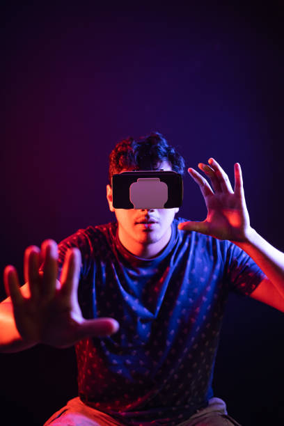 istock Young latin man using Virtual Reality VR Headset 3D Glasses, extends his hands to hold something. Dark backdrop. Low key 1389219714