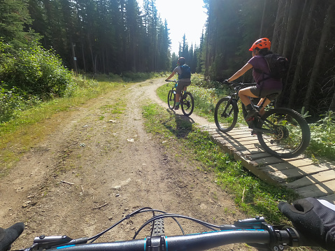 Personal perspective - adult daughter in 20s and mother in 50s cycling on dirt road in forest while father follows behind.  Sun Peaks, British Columbia, Canada.