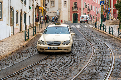 Lisbon taxi driving down cobblestoned hill in the city. There are people pictured walking on the sidewalks.