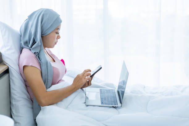 a asian women disease mammary cancer patient hold pick up smartphone casual working with laptop computer with pink ribbon wearing headscarf on bed in the bedroom at the house,healthcare,medicine - cancer cell cancer breast cancer breast imagens e fotografias de stock