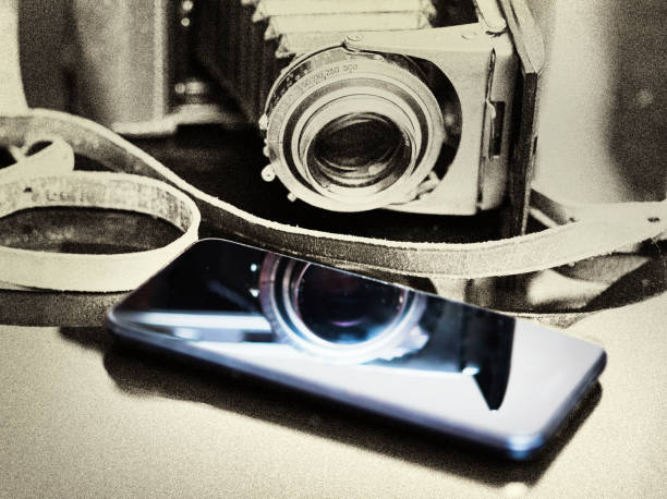 Progress in photography represented by an old-fashioned rangefinder camera paired with a modern mobile phone, the photographic tool of choice in the modern world. The scene is rendered in grainy sepia-toned black and white except for the cellphone, which shines out in modern color.