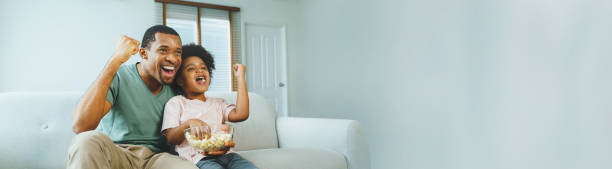 Cheerful African American Father and his boy watching and cheering sports games competition Cheerful African American Father and his little boy watching and cheering sports games competition together at home, Banner, Panoramic, celebrate excited football fan cheering for his team on tv stock pictures, royalty-free photos & images