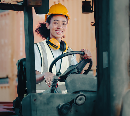 Smiling female foreman is driving forklift at import export container yard, portrait. African American industrial engineer woman drives reach stacker truck to load cargo box at logistic terminal port.