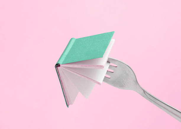 istock Minimal abstract composition made of fork holding a green book on isolated pastel pink background with copy space. Note card for book fair, promotions or education. Concept of reading or learning. 1389278973