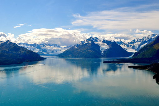 Aerial shot of the stunning Prince William Sound. Mountains and glaciers surround the beautiful azure waters of the bay, a small boat speeds along in the distance. Kenai Peninsula, Alaska.