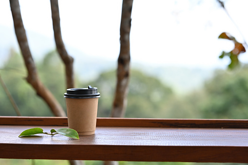A cup of take away coffee placed on the wooden balcony edge with natural background.