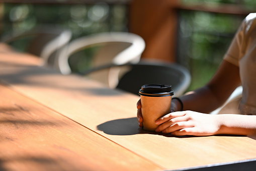 Close up with young female adolescence holding take away coffee cup among sunlight while sitting at wooden table.
