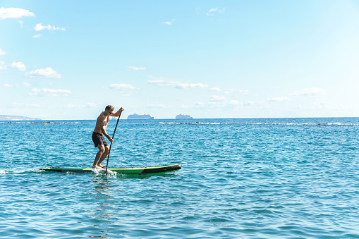 Young male surfer is riding a standup paddleboard and rowing with a paddle in ocean