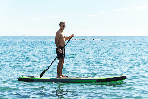 Young male surfer is riding a standup paddleboard and rowing with a paddle in ocean.