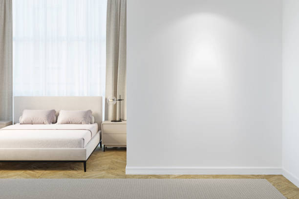A light bedroom with a blank illuminated white wall, a modern black lamp on the bedside table, a beige bed with light linen near curtained window, light beige carpet on the parquet floor.