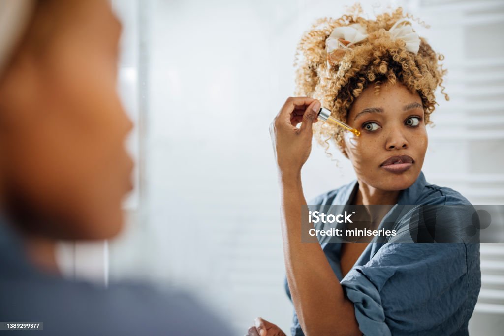 Morning routine: Woman Applying Face Serum Around her Eyes Beautiful African-American woman holding a face serum pipe and applying it around her eye. She is looking at herself in the mirror. Face Serum Stock Photo