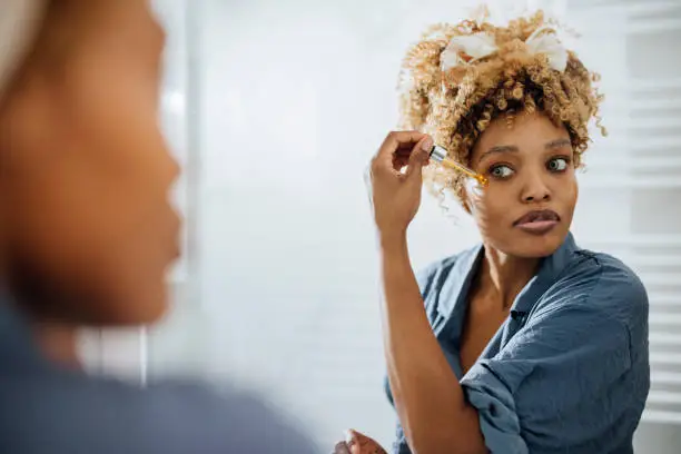 Beautiful African-American woman holding a face serum pipe and applying it around her eye. She is looking at herself in the mirror.