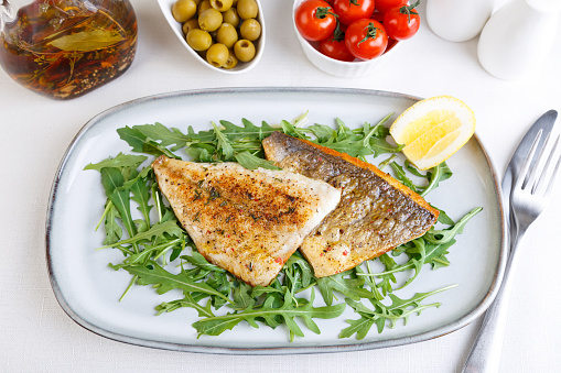 Fried dorado fillet with crispy skin. Arugula, cherry tomatoes, olives, lemon and olive oil. A traditional Mediterranean dish. White background. Selective focus, close-up, top angle.