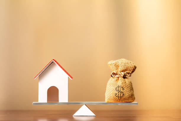 A scale weighing a miniature house and a little pocket of money