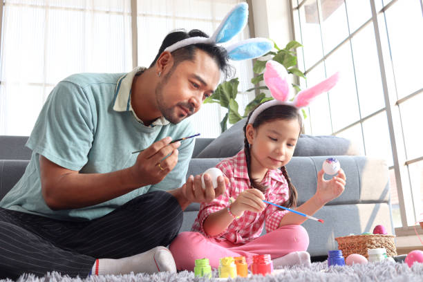 padre e hija preparando hermosos huevos de pascua para la decoración y celebración del comienzo de la primavera, adorable niña conejita rosa y su padre con diadema de orejas de conejo pintando huevos de colores juntos. - disfraz de conejo fotografías e imágenes de stock