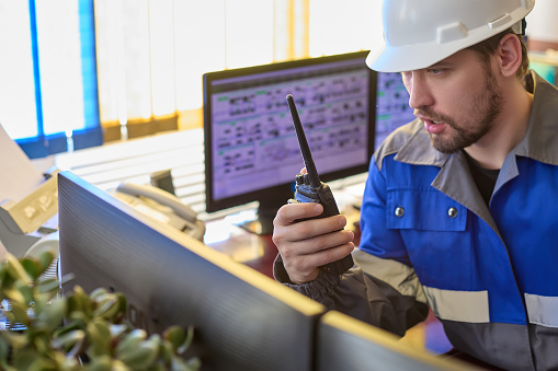 Serious Caucasian unshaven worker in overalls and helmet uses a walkie - talkie sitting in the control room . Automated workplace of a dispatcher or operator of a modern enterprise