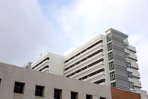 A low-down view, a close-up view of the white concrete building of a large old hospital with many floors and clouds in the sky as a backdrop, common in Thai towns.