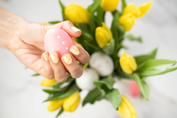 Woman with yellow manicure holding an Easter egg, bunch of tulips in the background.