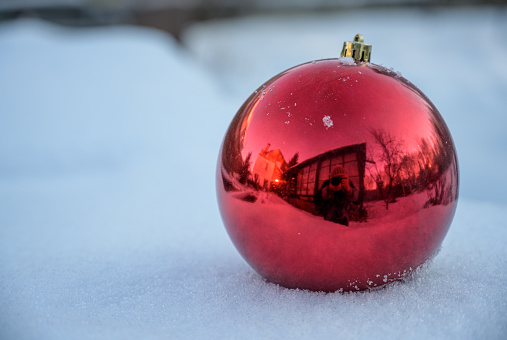 Glass big red ball - decoration for the New Year tree and interior. The Christmas tree toy lies in the snow, all the objects around it are reflected in it