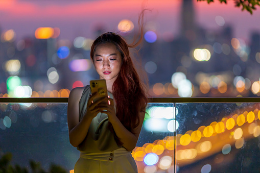 Asian woman using mobile phone with Bangkok skyscraper cityscape at sunset blue hour at downtown urban district for modern luxury living and prestige travel