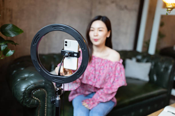 istock Smiling Asian Woman streaming broadcast In modern Cafeteria and using ring lamp and mobile phone 1389326121