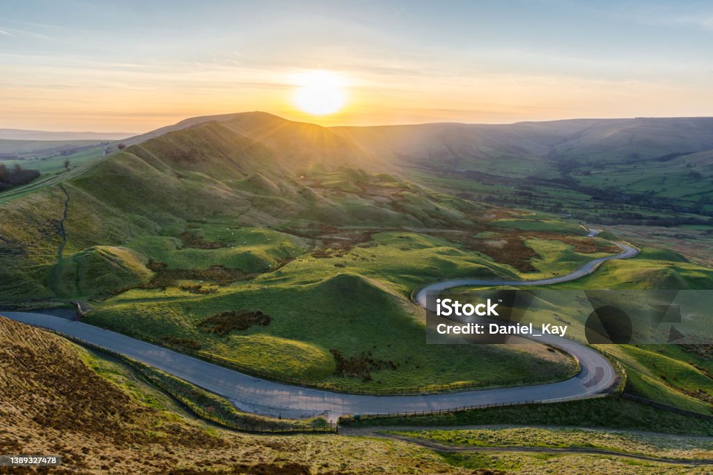 Sunset at Mam Tor in the Peak District with long winding road leading through valley. A long and winding rural road leading through lush green British countryside on a beautiful Summer evening. Taken from Mam Tor in the Peak District, UK. Journey Stock Photo