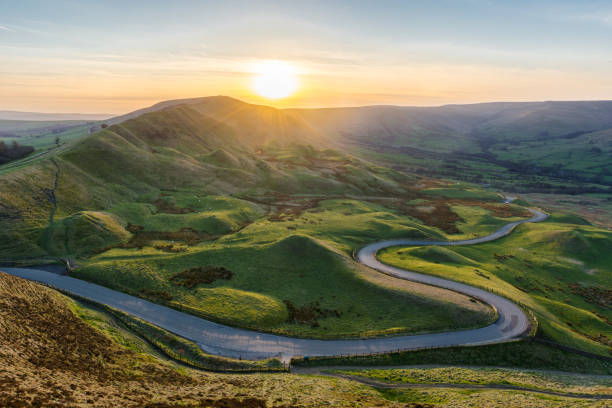 Sunset at Mam Tor in the Peak District with long winding road leading through valley. A long and winding rural road leading through lush green British countryside on a beautiful Summer evening. Taken from Mam Tor in the Peak District, UK. progress stock pictures, royalty-free photos & images