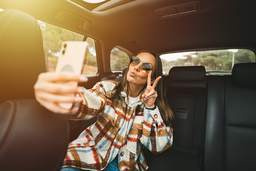 Stylish young woman sitting on back seat in the car and making selfie with smartphone. Woman taking picture with phone while sitting in taxi. High quality photo