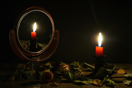 A burning candle reflected in a mirror standing on a wooden table among withered roses.