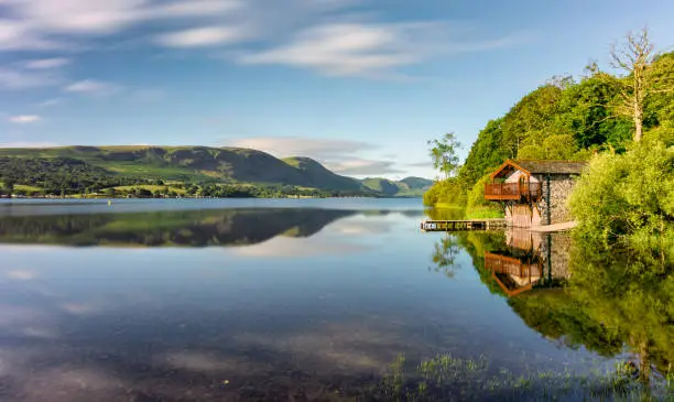 The iconic Duke of Portland Boathouse on the shoreline of Ullswater in the Lake District, UK. Taken on a sunny morning, a few clouds can be seen with a long exposure effect in the sky.