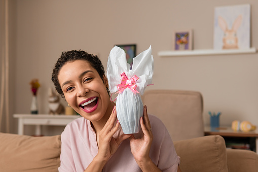 Happy woman showing white easter egg with pink bow at home.