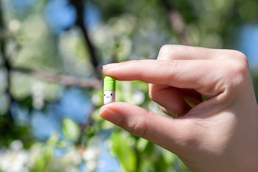 Woman holding in his hand a funny smiling pill among flowering trees in the garden. Seasonal Allergy. Spring time. Creative