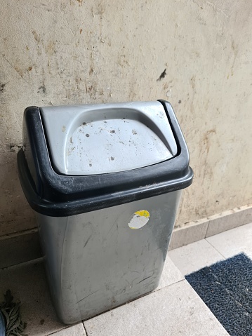 close up of a very dirty gray plastic trash can on the tiled floor against a dirty and dusty wall background