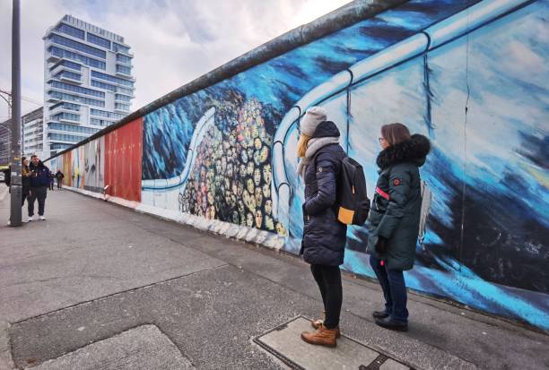 Berlin, Germany - May 30, 2025: Wide-angle view of the iconic Berlin Wall covered in vibrant graffiti, contrasting with modern buildings in the Friedrichshain district under a bright sky.