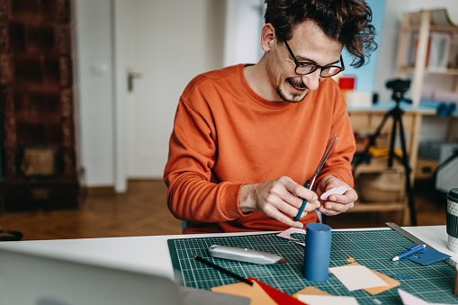 Smiling male design professional cutting papers with scissors on desk while sitting in creative studio