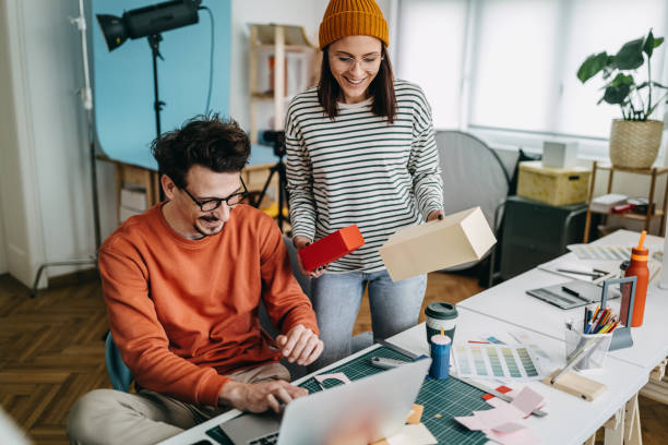 Woman holding boxes while male coworker using laptop in office