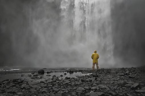 Man wearing yellow raincoat enjoying scenic view to huge and powerful Skogafoss waterfall in South Iceland