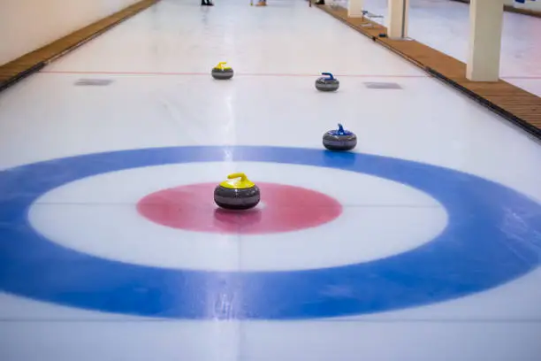 A yellow-handled curling stone sits on the ice in the center of the house, with the other stones out of focus in the back.