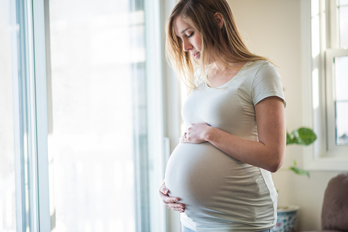 Beautiful, bright side view of a pregnant woman in a light shirt holding her stomach tenderly with a neutral expression on her face as she is looking out the window.