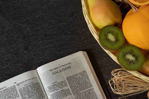 An open Holy Bible book of Galatians with a wicker basket full of various fresh fruits with copy space. The biblical concept of the Christian fruit of the Spirit. Love, joy, peace. Top view.