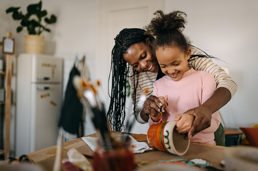 Happy mother with dreadlocks holding daughter's hands and assisting her in painting flower pot with watercolor paints at home