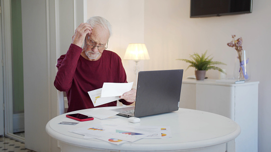 An elderly man pays taxes via the Internet. He scratches his head thoughtfully and examines bills